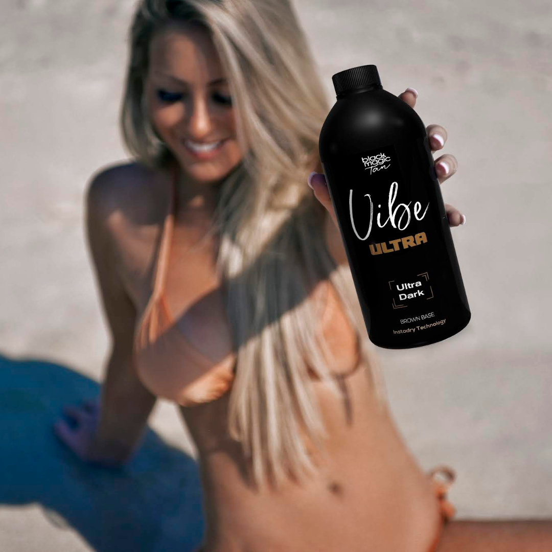 Woman on the beach holding a bottle of Vibe Ultra spray tan solution, with a deep bronzed glow and blonde hair in the sunlight.