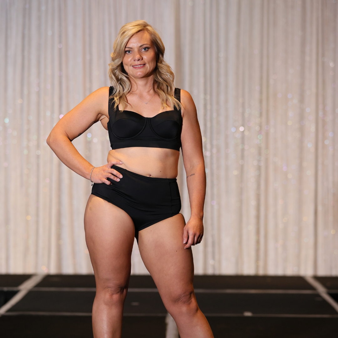 Woman in a black bikini posing on a runway with a deep, violet-base spray tan finish.